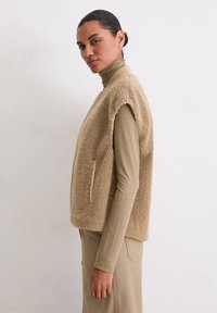 Beige Fleece-Weste mit weicher Textur, ärmellosem Design und Seitentaschen, getragen über einem enganliegenden hellbraunen Oberteil.