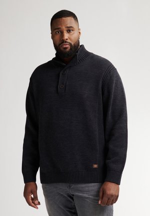 Hombre con barba que lleva un suéter de punto oscuro abotonado y jeans grises, de pie frente a un fondo claro y liso.