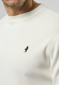 Witte sweater van wafelstructuur met een ronde hals en een klein zwart geborduurd logo op de borst. Het materiaal lijkt textured en ademend.