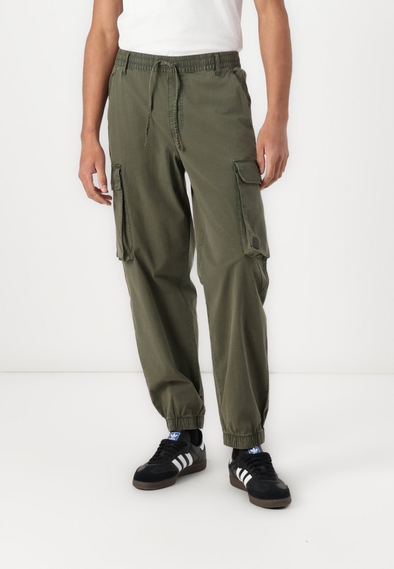 Olivgrüne Cargo-Hosen mit elastischem Bund, Kordelzug und zwei seitlichen Taschen. Der Stoff sieht leicht aus und hat eine glatte Textur.