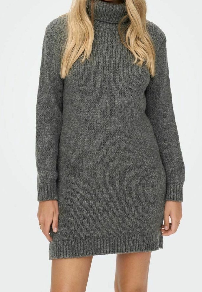 Femme portant une robe-pull en tricot gris à col roulé et manches longues, les mains détendues le long du corps sur un fond uni.