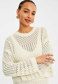 Sweter w kolorze kremowym, wykonany z dzianiny, z luźnym splotem, prążkowanym dekoltem i mankietami, o oversize'owym kroju oraz teksturowanych detalach.