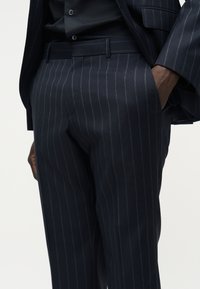 Pantalon de costume bleu marine à fines rayures porté avec une veste assortie, main de la personne dans la poche droite, chemise noire rentrée, gros plan sur le bas du torse.