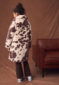 Przytulny płaszcz z beżowego i brązowego polarowego materiału w plamki, z szerokim kołnierzem, zestawiony z brązowymi szerokimi spodniami i jasnoniebieskimi tenisówkami. Widok od tyłu.