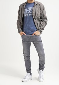 Hombre que lleva una chaqueta gris, una camiseta gráfica azul, pantalones vaqueros grises y zapatillas blancas, de pie con las manos en los bolsillos frente a un fondo simple.