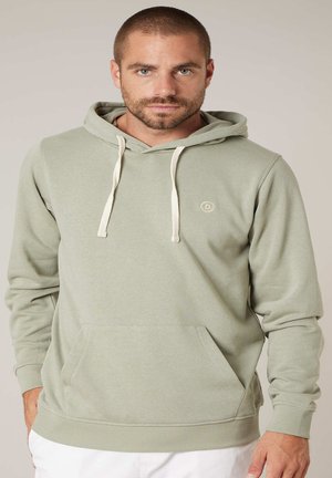 Man met kort haar en baard, gekleed in een lichtgroene capuchontrui en witte broek, staand tegen een neutrale achtergrond.