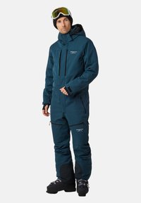 Teal overall ski pak met een capuchon, rits aan de voorkant, borstzak en zijzakken met rits. Voorzien van versterkte kniepanelen en een gladde textuur.