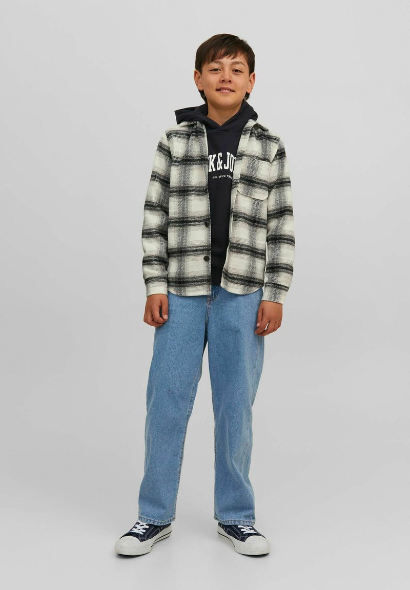 Camisa de cuadros en blanco y negro sobre un hoodie azul marino, jeans de mezclilla azul claro y zapatillas azul marino. Diseño casual con botones en el frente y cuello abierto.