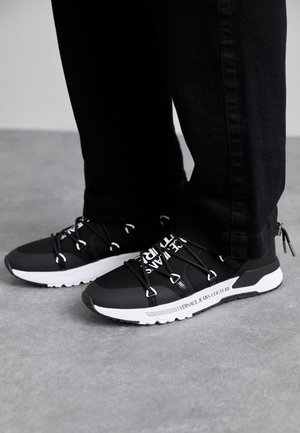 Baskets de sport noires avec une semelle et des détails blancs contrastants. Présente des lacets élastiques et un logo sur le côté. Portées avec un jean noir.