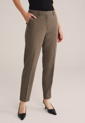 Vrouw draagt taupekleurige rechte broek met voorkeerplooi, hand in de zak, gecombineerd met zwarte slingback hakken met puntige neus en zwarte top.