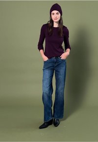 Donkerpaarse lange mouwen top, losse blauwe spijkerbroek en zwarte enkellaarsjes. Het model draagt een bijpassende donkerpaarse gebreide beanie.