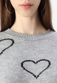 Grå strikket genser med ribbestrikket halskant og to svarte hjerteomriss. Stoffet ser mykt ut med en teksturert overflate.