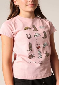 Roze katoenen T-shirt met een aansluitend ontwerp, versierd met verschillende geïllustreerde cowboylaarzen en hoeden. Onder aan staat de tekst "Het Meisje uit Texas."