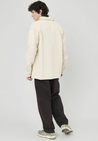 Veste chemise texturée beige avec un col, portée sur un pantalon marron foncé et des baskets grises ; vue de derrière.