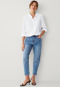 Camicia bianca in lino con bottoni frontali e maniche arrotolate. Jeans azzurri dritti con orlo sfilacciato. Sandali neri con tacco. Sfondo semplice.