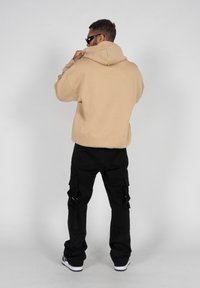 Haut à capuche surdimensionné beige avec une grande capuche, associé à un pantalon cargo noir avec des poches latérales, porté avec des baskets blanches. L'arrière-plan est uni.