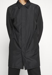 Black, lightweight, long coat with a pointed collar, front button closure, and a smooth, shiny texture. Features two side pockets.