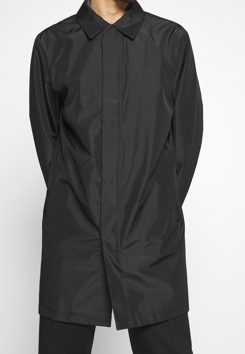 Black, lightweight, long coat with a pointed collar, front button closure, and a smooth, shiny texture. Features two side pockets.