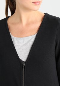 Haut noir à fermeture éclair avec tissu texturé, doté d'un col en V et d'une chemise grise superposée en dessous. Détails de la fermeture éclair visibles.