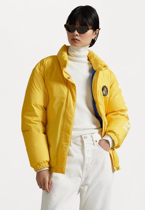 Chaqueta de plumas amarilla con interior azul, cierre de botones a presión, cuello alto, detalles en blanco y logo. Llevada sobre un suéter de cuello alto blanco.
