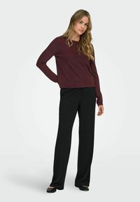 Suéter burdeos de manga larga con un escote redondo, combinado con pantalones anchos negros yloafers negros, sobre un fondo liso.