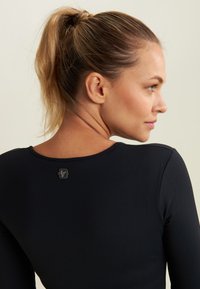 Top nero a maniche lunghe con scollo rotondo; texture liscia e piccolo logo sul retro del collo. Pelle visibile al collo.