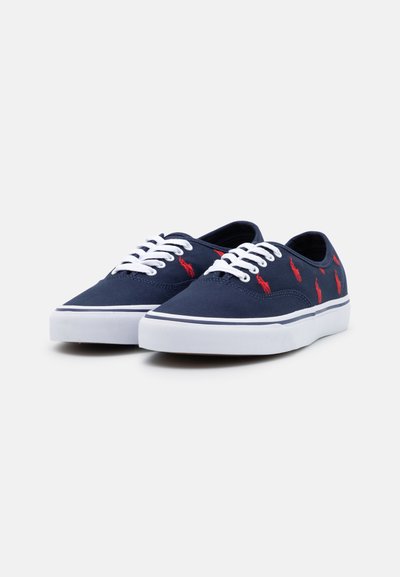 Sapatilhas de lona azul-marinho com atacadores brancos e solas de borracha, apresentando logótipos bordados em vermelho. Forma arredondada e textura suave.