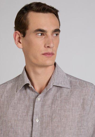 Joven con cabello castaño que lleva una camisa de botones de rayas marrón claro, mirando hacia su derecha contra un fondo gris sencillo.