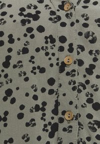 Camicia button-up in tessuto grigio chiaro con un motivo a macchie nere. Dettagli notevoli includono bottoni tondi color cammello e una texture sottile.
