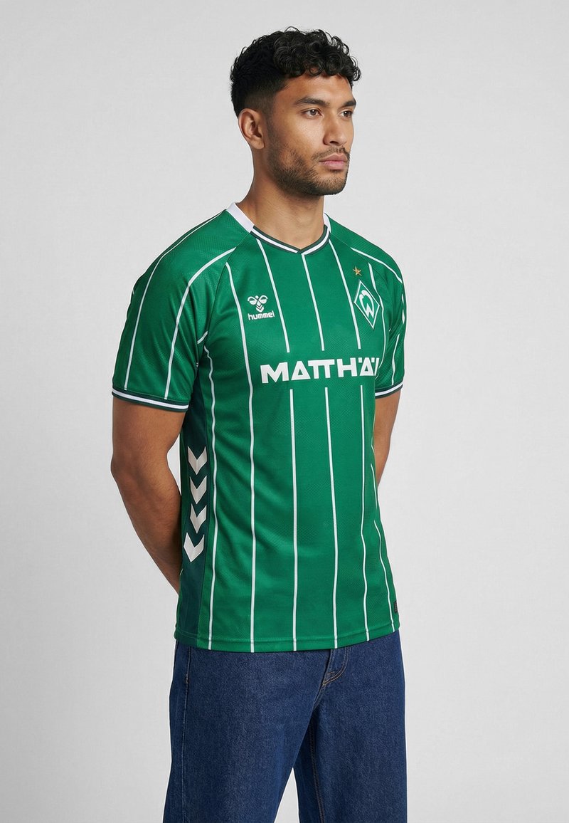 Mees kannab rohelist Werder Bremeni jalgpallimängija särki valgete triipude ja logoga, seisab käed selja taga tavalise tausta ees.