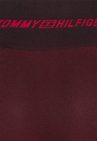Tommy Hilfiger ribbed tights in dark burgundy with a wide black waistband featuring a bold red logo. Smooth texture and form-fitting design.