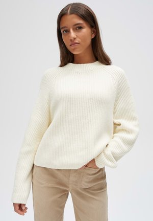 Jonge vrouw met steil bruin haar, gekleed in een crèmekleurige gebreide trui en beige broek tegen een effen achtergrond