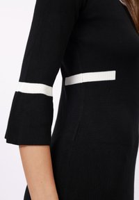 Black knitted dress with three-quarter sleeves. White horizontal stripes at sleeve hems and a side detail. Ribbed texture throughout.