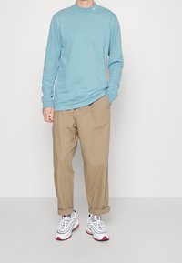 Man som bär en ljusblå långärmad skjorta, beige uppvikta byxor och vita sneakers med röda och grå accenter, står mot en enfärgad bakgrund.