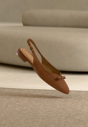 Brun ruskindslå på flad sko med en lille sløjfe på tåen og guldlås, vist mod en beige sofa og tæppe.