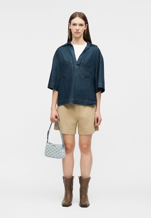 Woman wearing loose dark denim shirt, beige shorts, brown ankle boots, holding small blue patterned handbag against plain background.