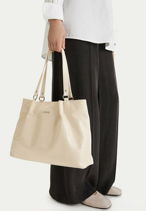 Mano che tiene una grande borsa tote beige con trama, dotata di doppi manici, indossata con pantaloni neri a gamba larga, camicia bianca e scarpe beige piatte.