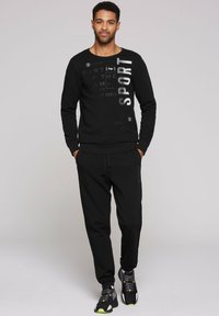 Sudadera negra con texto gráfico y pantalones de chándal negros a juego. El conjunto tiene un corte relajado y se complementa con zapatillas negras con acentos amarillos.