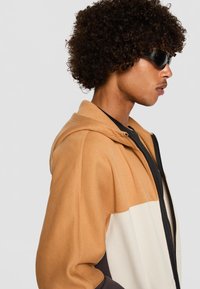 Sudadera con capucha con diseño bicolor en marrón, beige y crema. Confeccionada en una tela suave, cuenta con cierre de cremallera y bolsillos laterales.