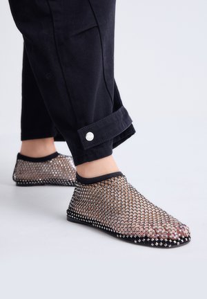 Pies con zapatos negros de malla sin cordones con recortes en forma de diamante, combinados con pantalones negros cortos con puño abotonado.