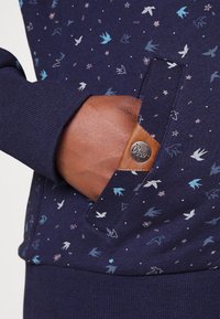 Sudadera navy con un patrón floral y de aves en varios colores, con un bolsillo texturizado y un acento de logo en cuero.