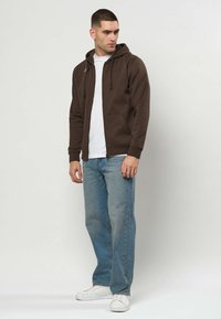Homme debout regardant sur le côté, portant un sweat à capuche marron zippé, un t-shirt blanc, un jean bleu et des baskets blanches sur un fond uni.