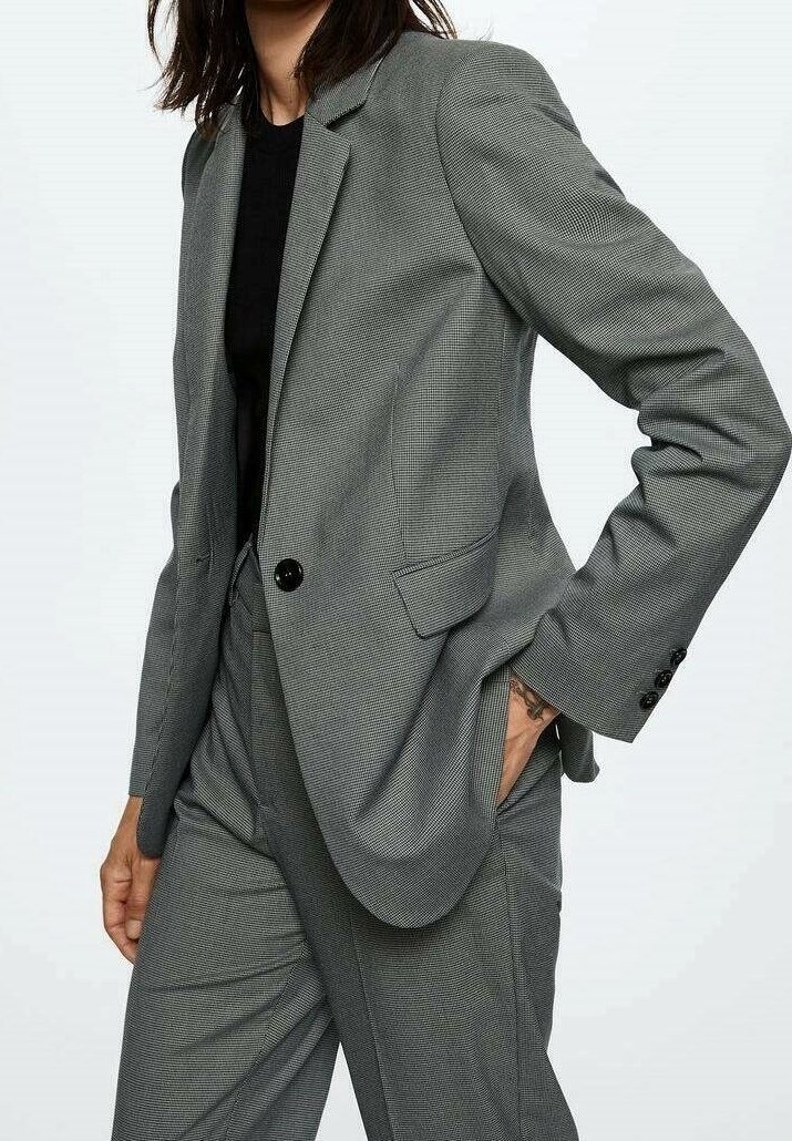 Femme portant un costume à carreaux gris avec une chemise noire, debout avec une main dans la poche sur un fond clair uni.