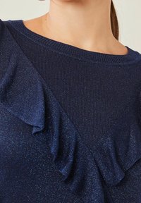 Pull en tricot bleu marine avec un tissu texturé, un col rond et un détail de volants diagonal sur le devant.
