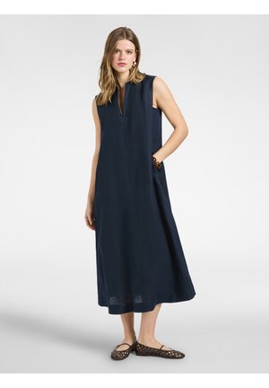 Frau trägt ärmelloses marineblaues Midikleid mit Seitentaschen und braunen gewebten flachen Schuhen, steht vor einem schlichten hellen Hintergrund.