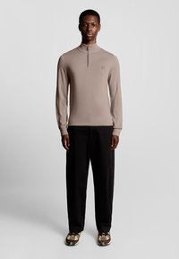 Beige Pullover mit Reißverschluss und weichem Strickmaterial, kombiniert mit schwarzen Hosen und gemusterten Schuhen. Schlichtes Design mit einem dezenten Logo-Detail.