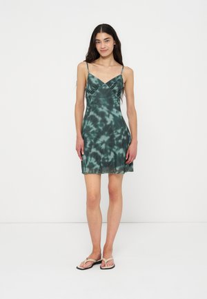 Femme debout, portant une robe verte sans manches à motif tie and dye avec des bretelles fines et des tongs blanches, posant devant un fond blanc uni.