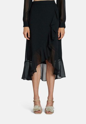 Jupe midi noire avec des volants superposés, en tissu transparent, et un ourlet évasé. Associée à des sandales beiges avec des embellissements floraux.