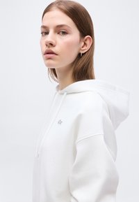 Jeune femme aux cheveux châtain clair portant un sweat à capuche blanc avec un petit logo crocodile sur la poitrine, debout devant un fond clair uni.