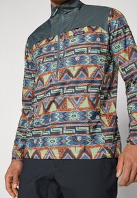 Kort zip-pullover i stof med geometriske mønstre i teal, brunt og grønt. Har en struktureret mørkegrå bærestykke. Glat finish generelt.